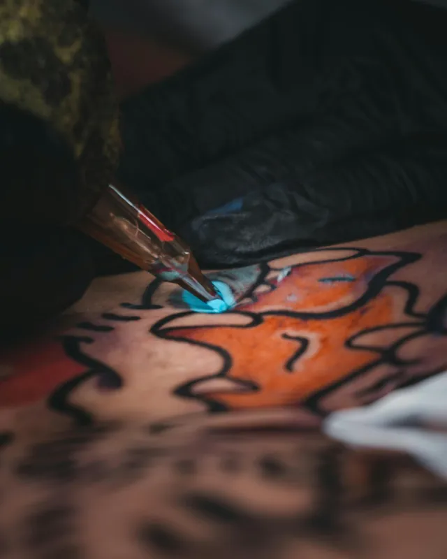 A tattoo needle packing orange and blue color into a bold outline