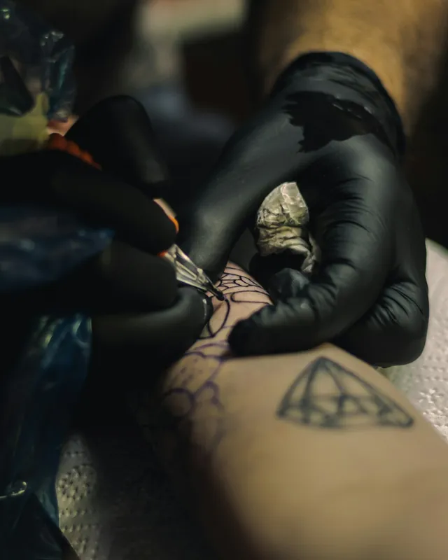 Gloved hands tattooing fine line work on a forearm beside a small geometric tattoo