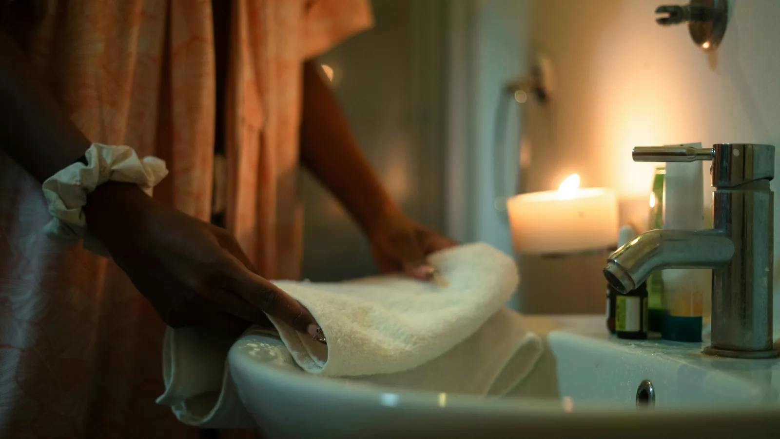 Hands folding a soft white towel beside a sink in warm candlelight