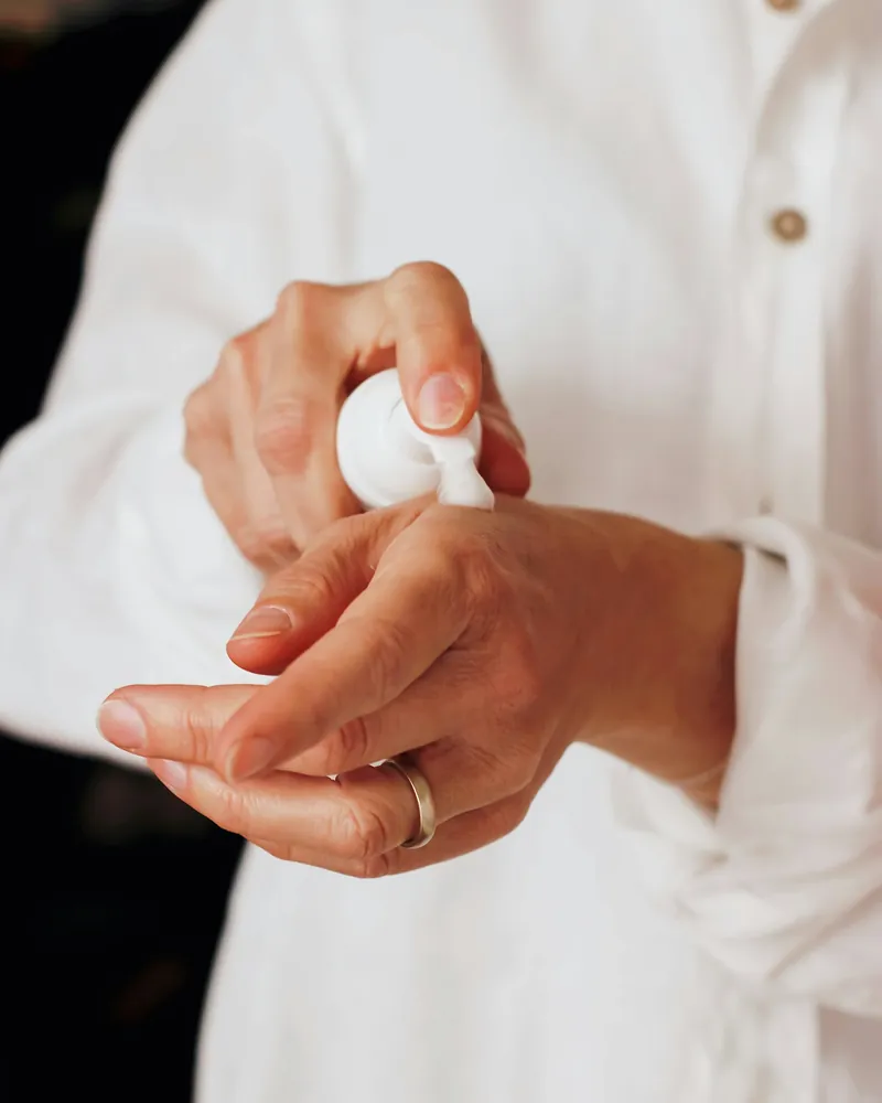 Hands in a white linen shirt pumping moisturizer from a white bottle